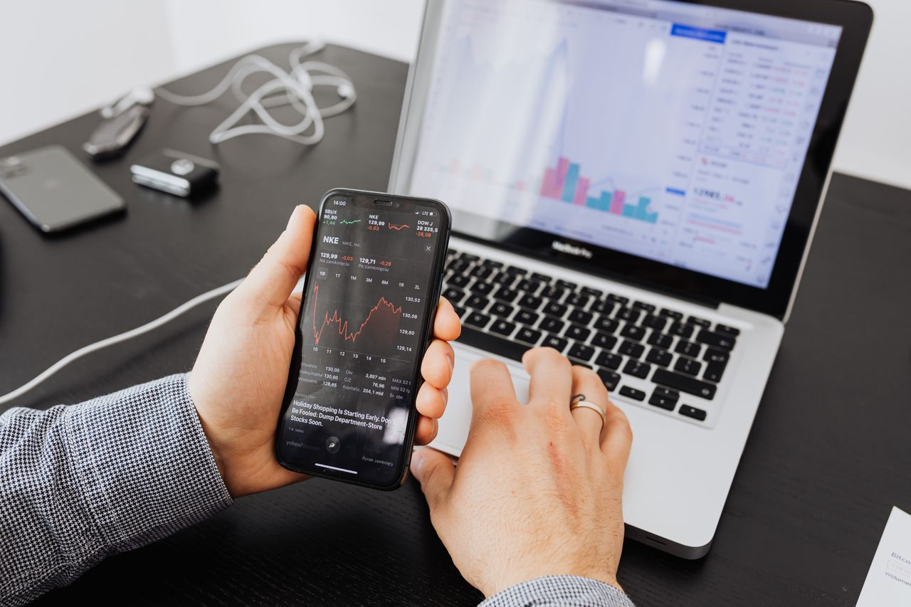 A person reviews stock data using a smartphone and laptop while indoors.